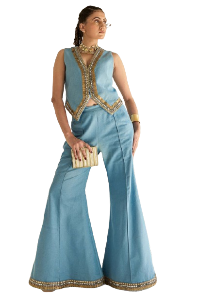 Woman in blue embellished vest and flared pants holding a clutch in a studio setting.